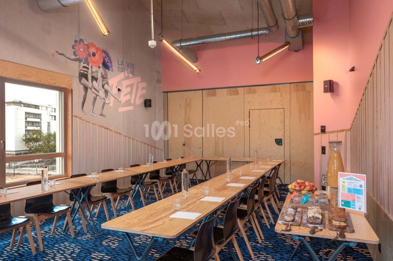 Salle de réunion lumineuse avec tables en bois, chaises noires, mur rose et buffet de collations sur le côté.