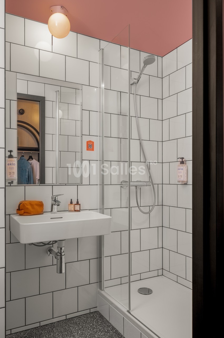 Salle de bain moderne avec douche vitrée, carrelage blanc, lavabo suspendu et éclairage mural.