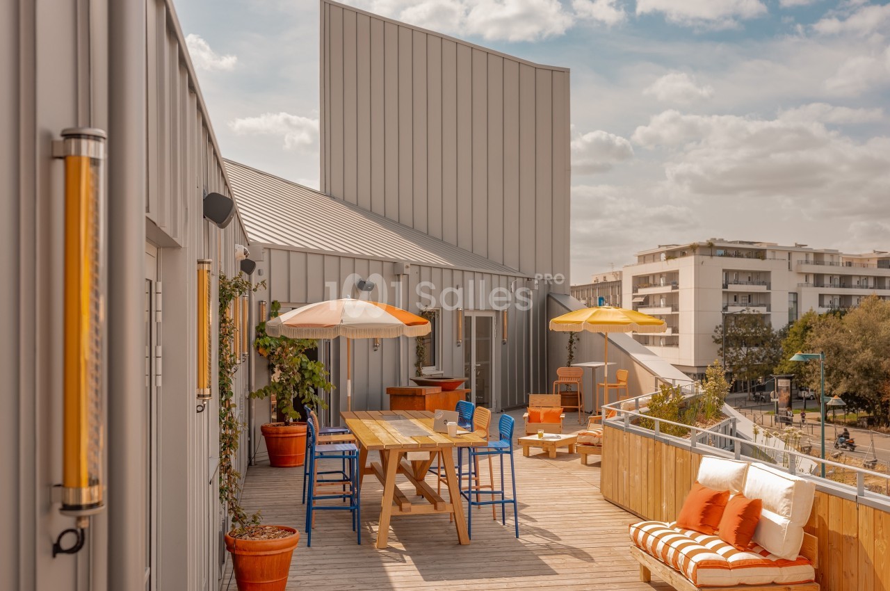 Terrasse en bois avec tables, chaises colorées, parasols, plantes en pot et vue sur des immeubles environnants.