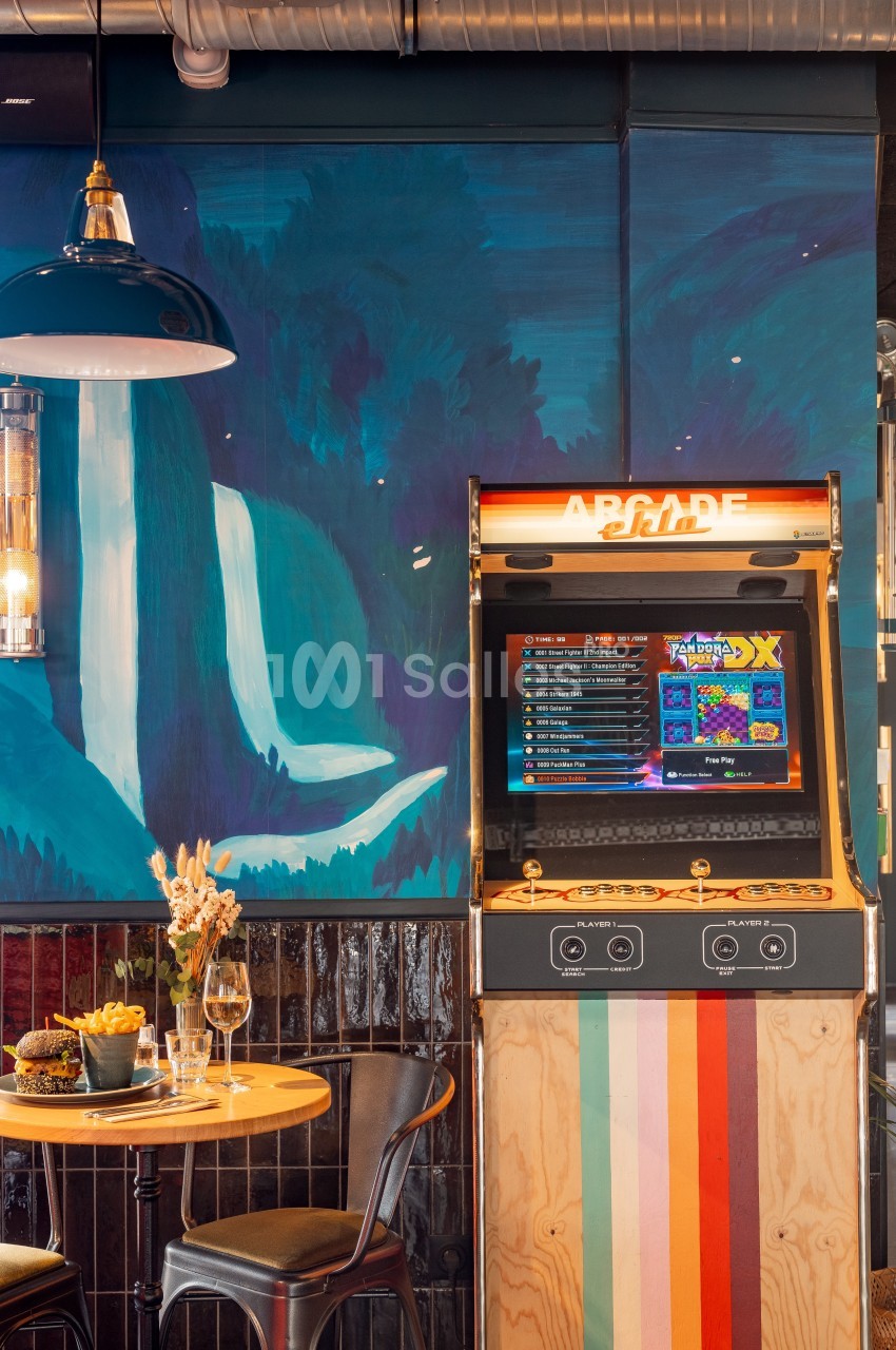 Borne d'arcade rétro près d'une table avec des boissons et des snacks, dans un décor coloré avec une fresque murale.