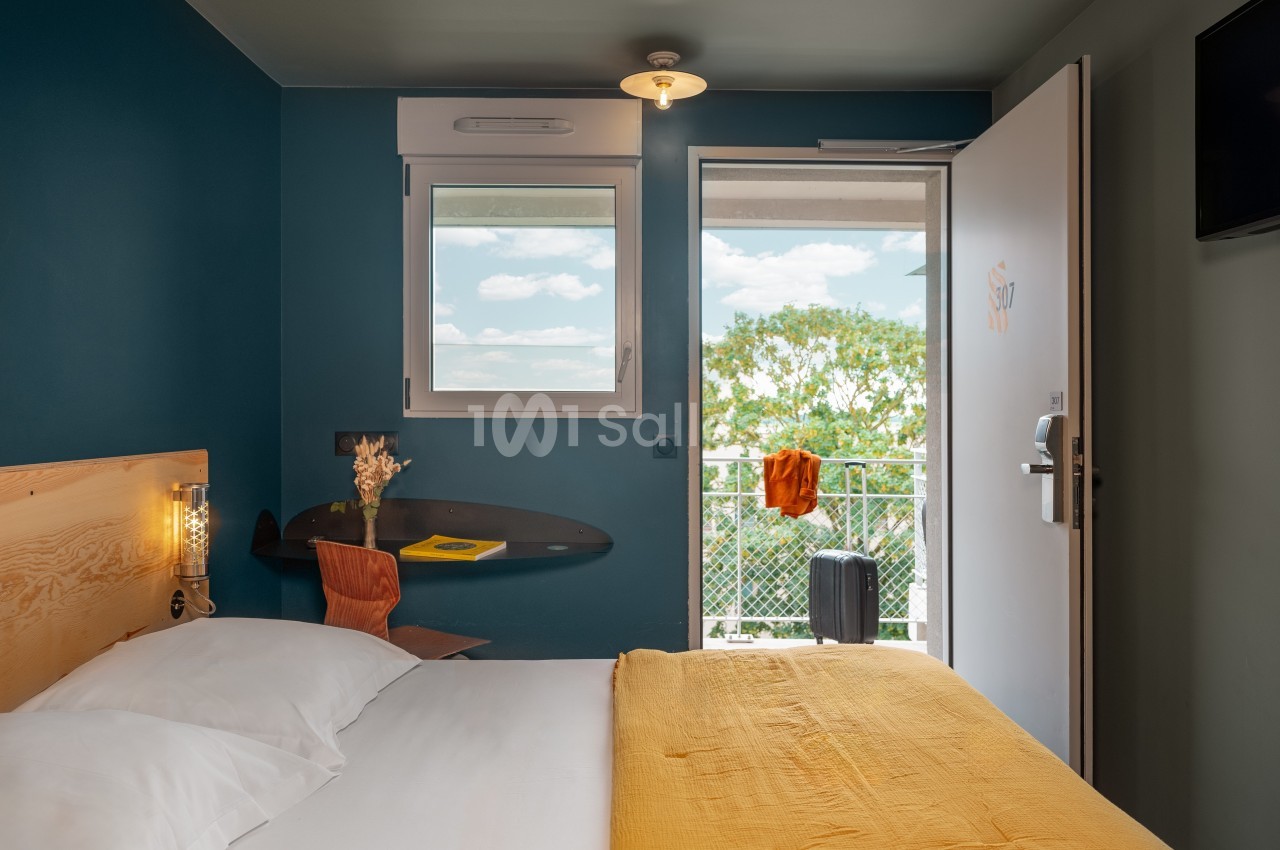 Chambre d'hôtel lumineuse avec lit double, bureau, fenêtre ouverte sur un balcon et valise visible à l'extérieur.