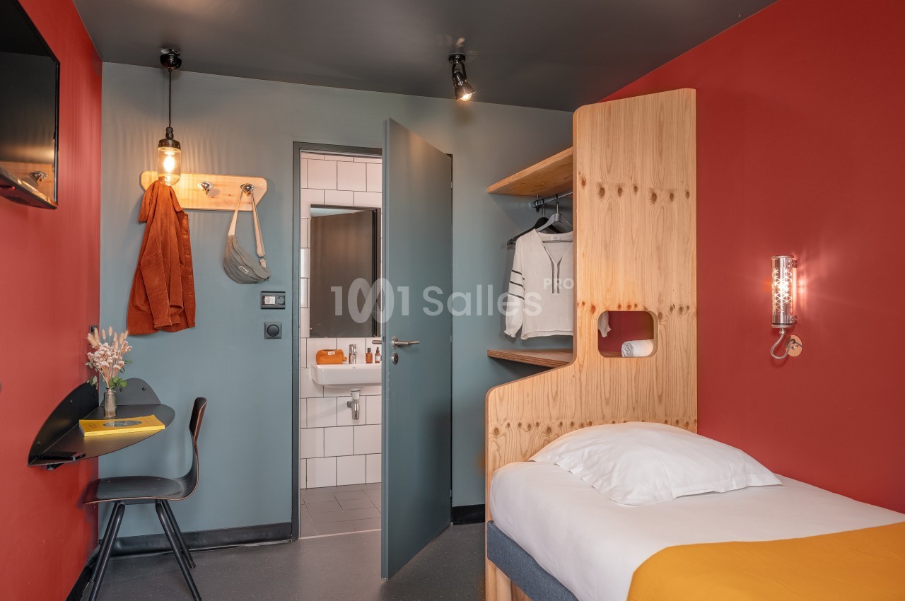 Chambre moderne avec lit simple, bureau, étagère en bois, et vue sur une salle de bain carrelée avec miroir.