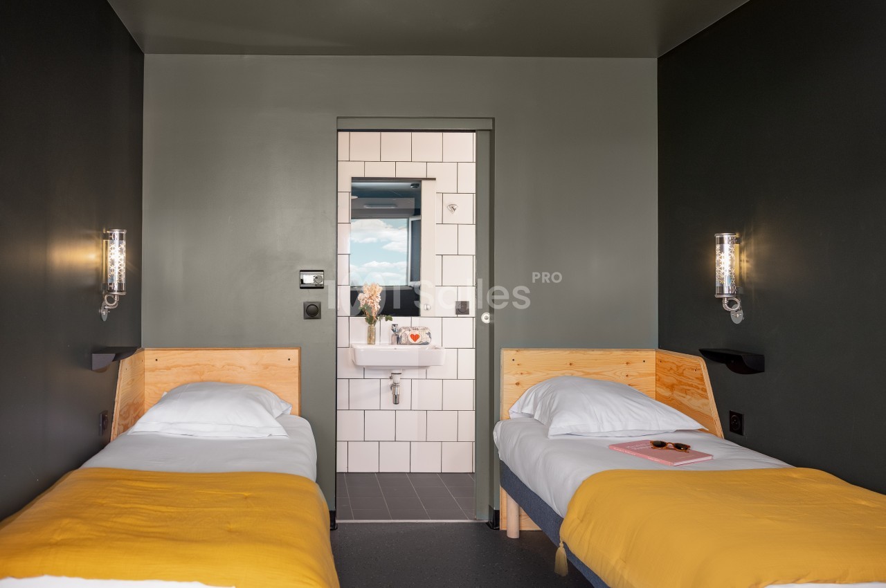 Chambre avec deux lits simples, draps jaunes, murs sombres et vue sur une salle de bain carrelée avec lavabo.