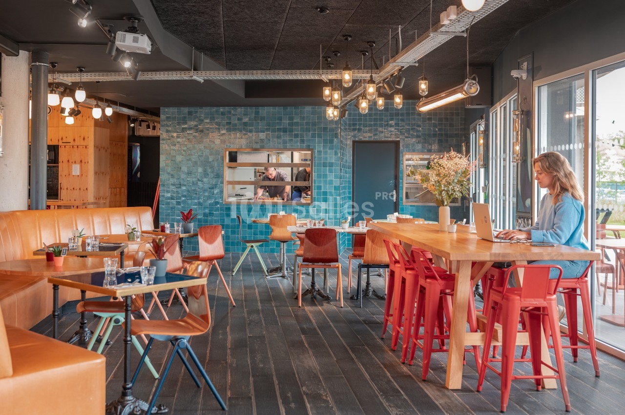 Espace lumineux avec tables en bois, chaises colorées, mur en carreaux bleus et une personne travaillant sur un ordinateur…