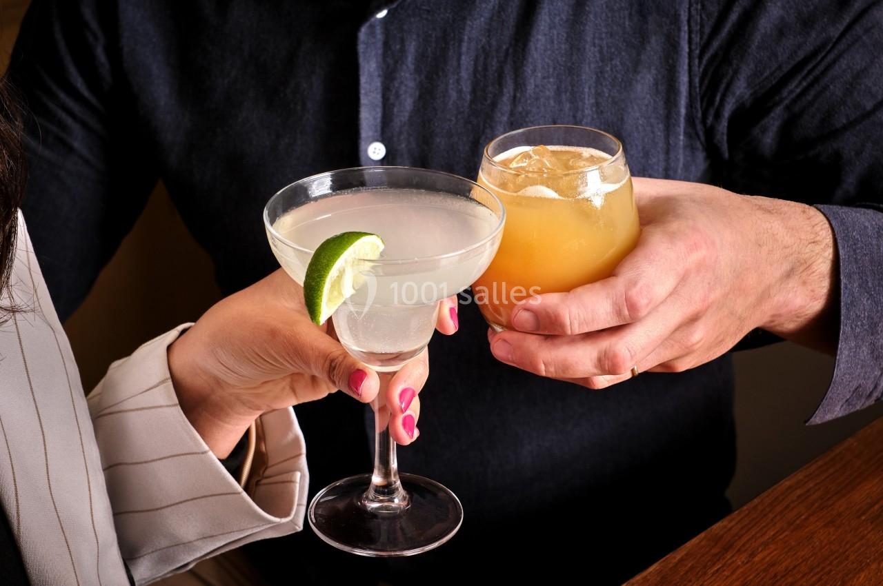 Deux personnes trinquent avec un cocktail clair garni d'une tranche de citron vert et une boisson ambrée sur glace.