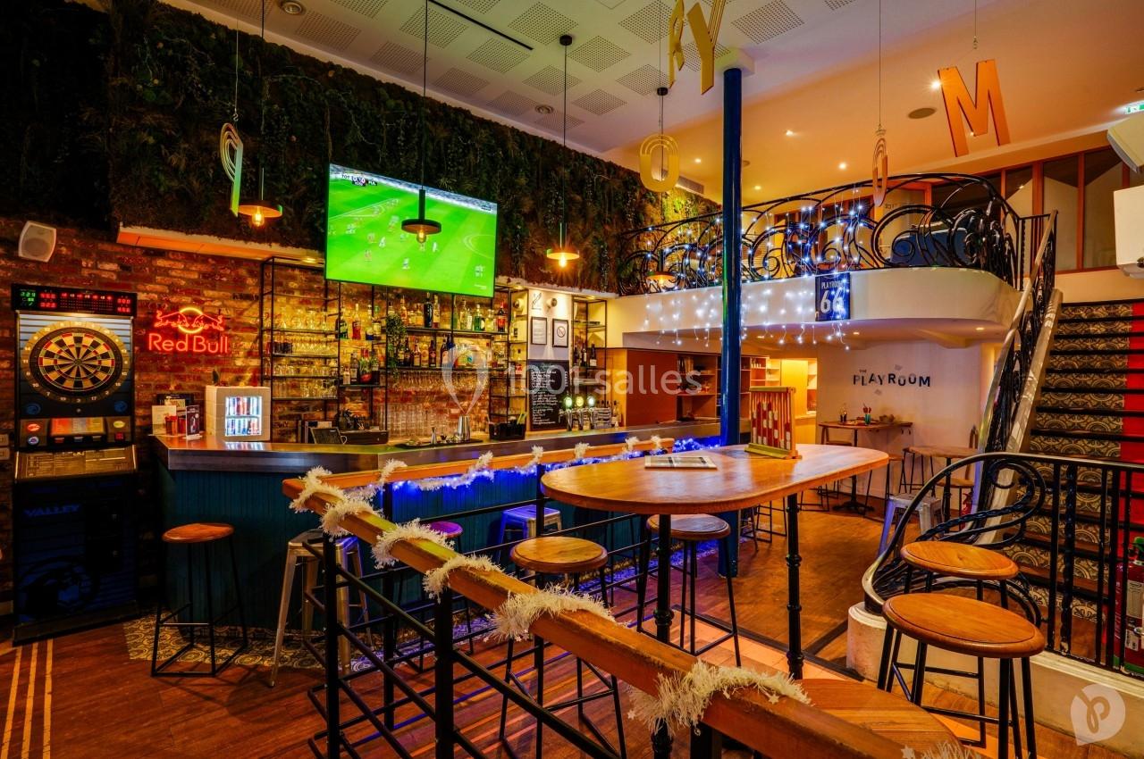 Bar avec comptoir éclairé, tables hautes en bois, écran diffusant un match, décorations lumineuses et mur végétal.