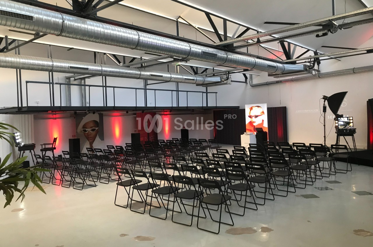Salle aménagée pour un événement avec des rangées de chaises noires, éclairage rouge et affiches publicitaires.