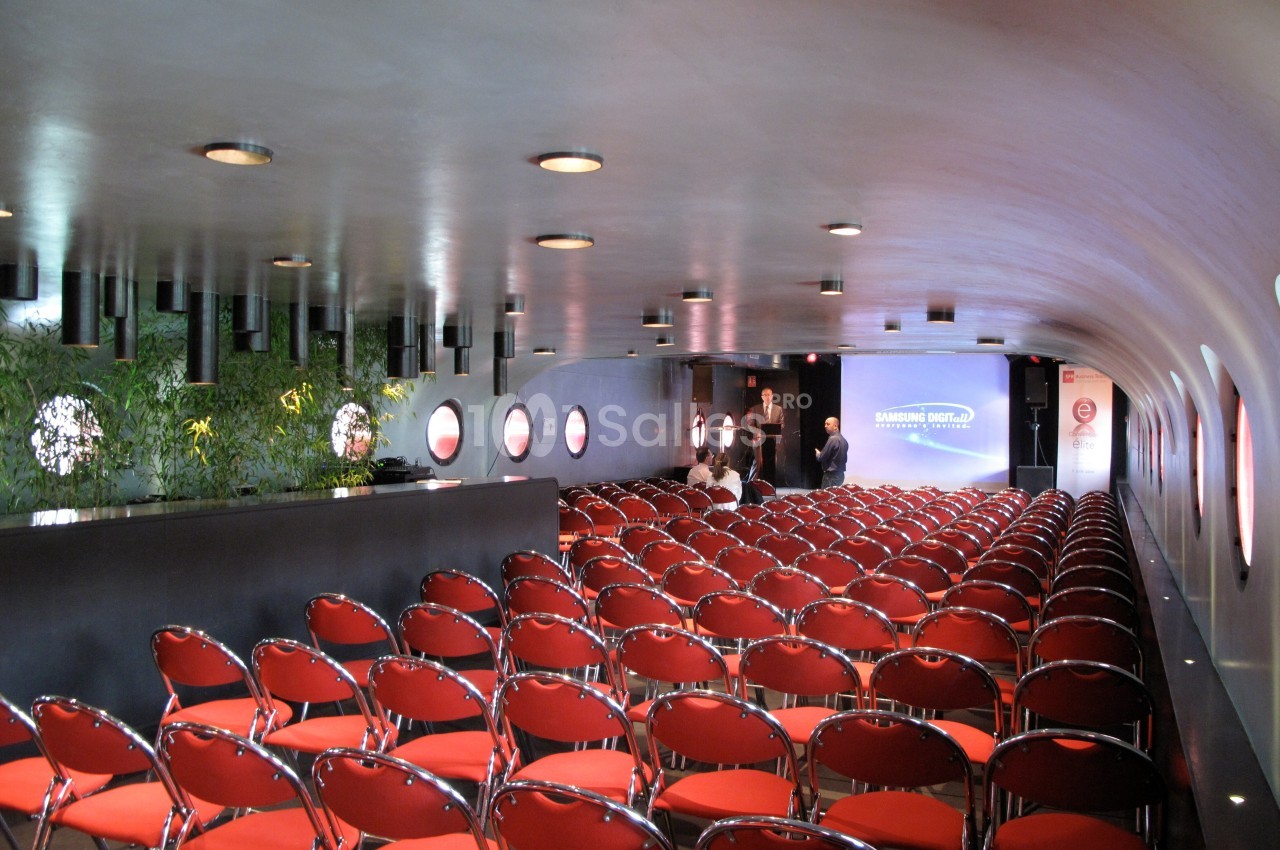 Salle de conférence moderne avec rangées de chaises rouges, éclairage tamisé et écran de présentation à l'arrière.
