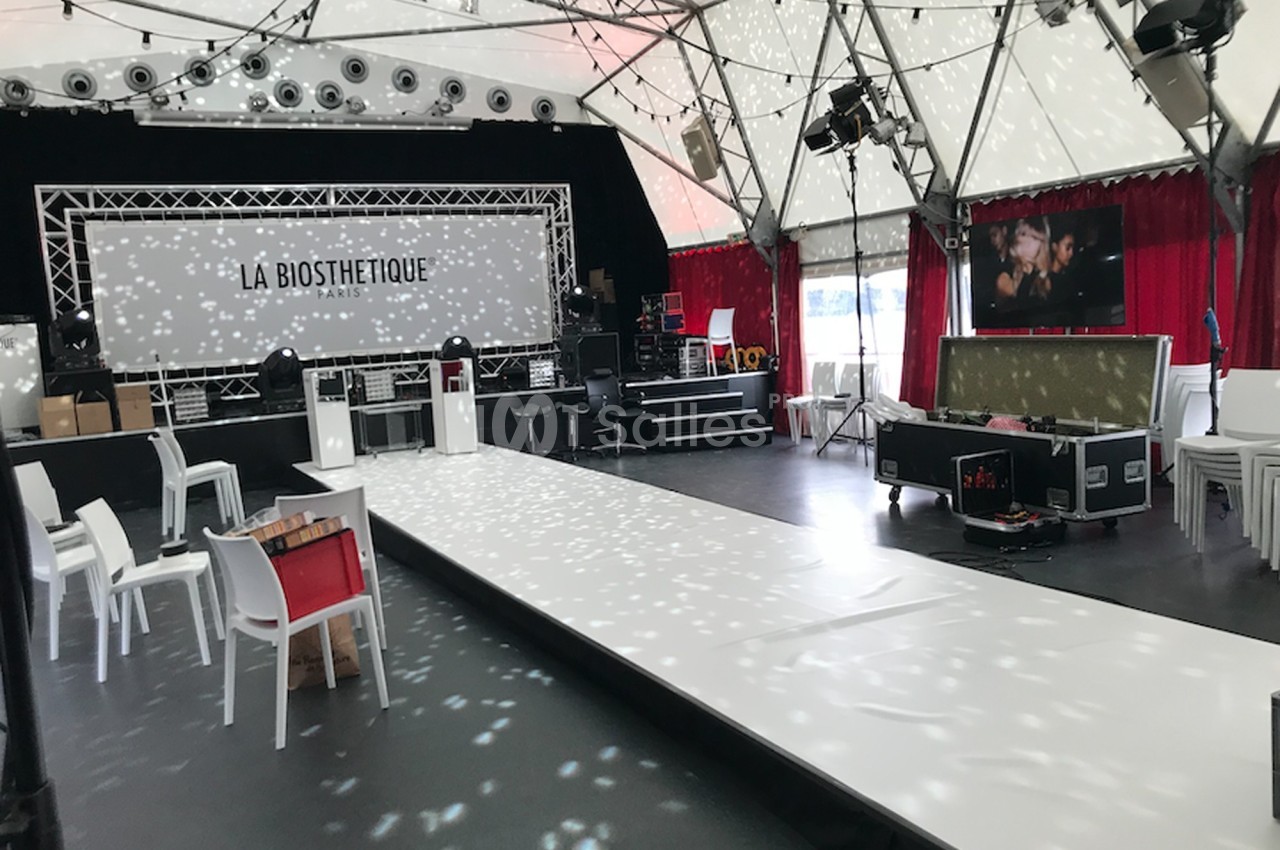 Scène et podium éclairés dans une salle sous chapiteau, avec chaises blanches et matériel technique installé.