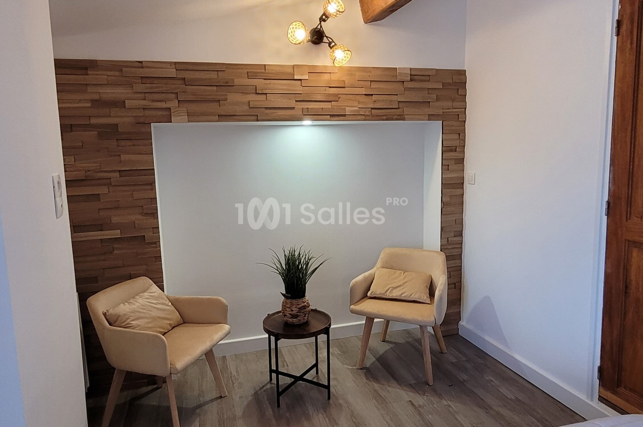 Deux fauteuils beige autour d'une table basse avec une plante, dans un coin décoré de bois et éclairé par une applique…