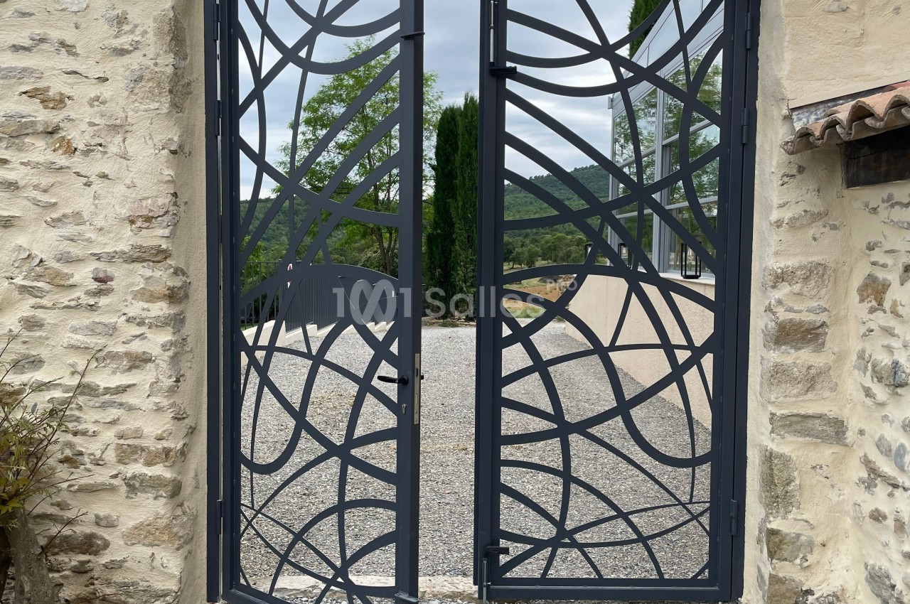 Portail métallique noir ajouré avec motifs abstraits, encadré par des murs en pierre, donnant sur une allée gravillonnée.