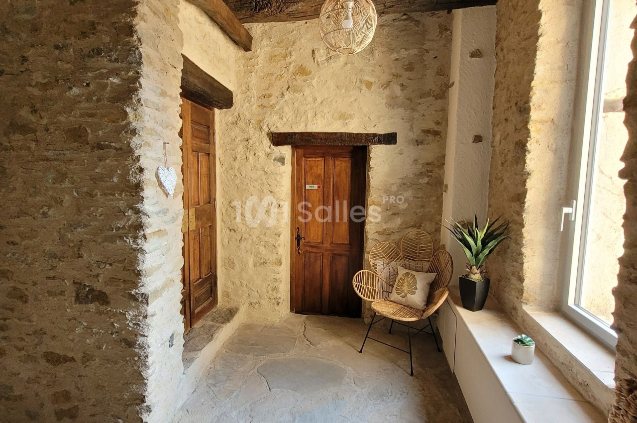 Espace intérieur en pierre avec porte en bois, fauteuil en osier, plante en pot et lumière naturelle provenant d'une fenêtre.