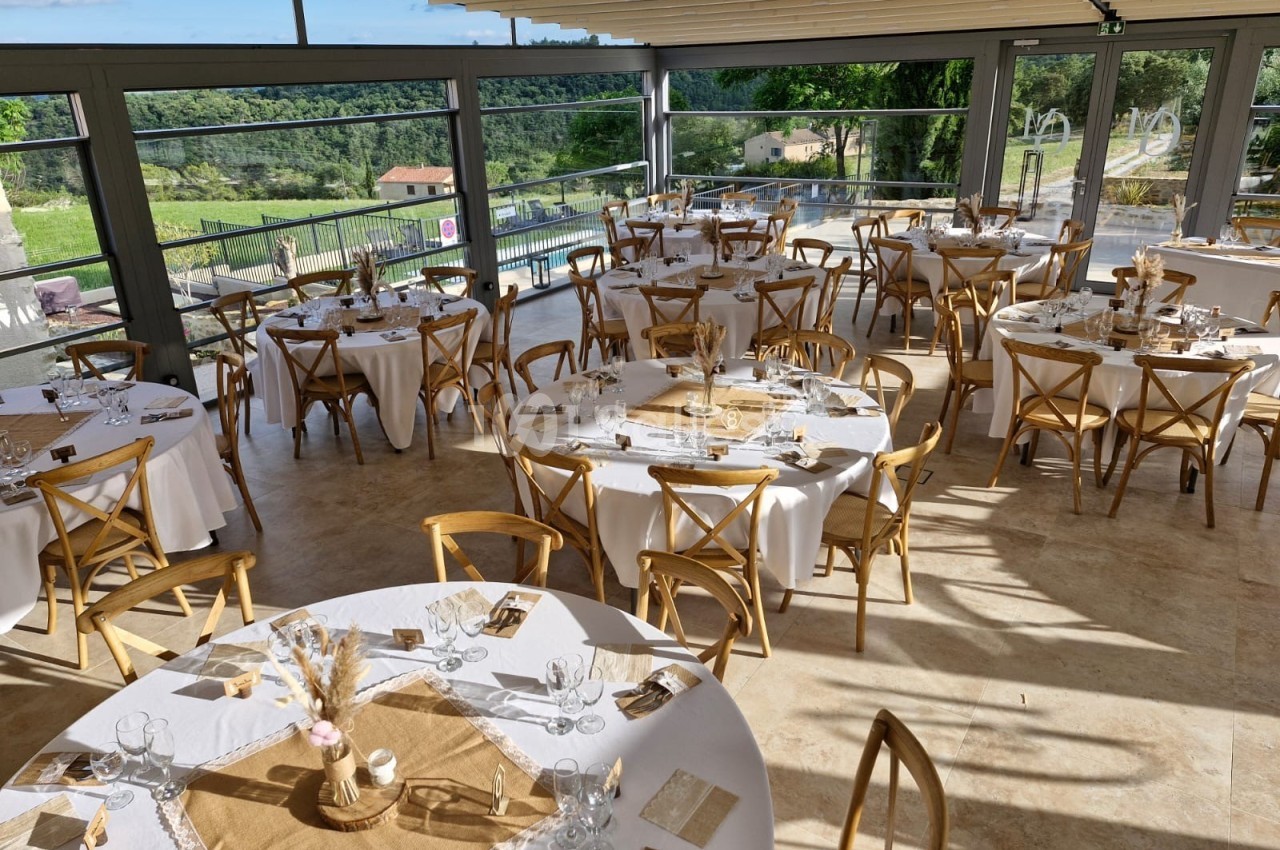 Salle lumineuse avec tables rondes dressées pour un événement, entourée de grandes baies vitrées donnant sur un paysage…