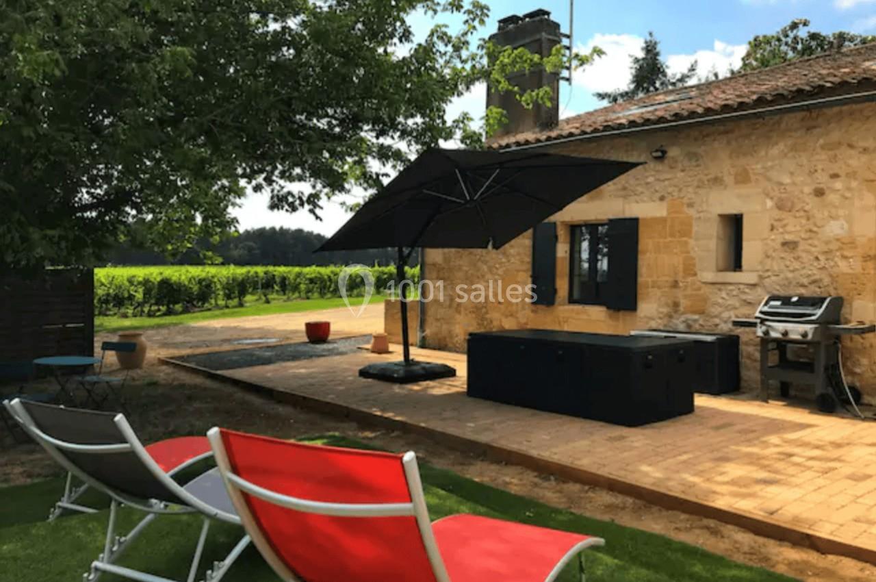 Terrasse en bois avec parasol, chaises longues colorées, barbecue et maison en pierre dans un cadre verdoyant.