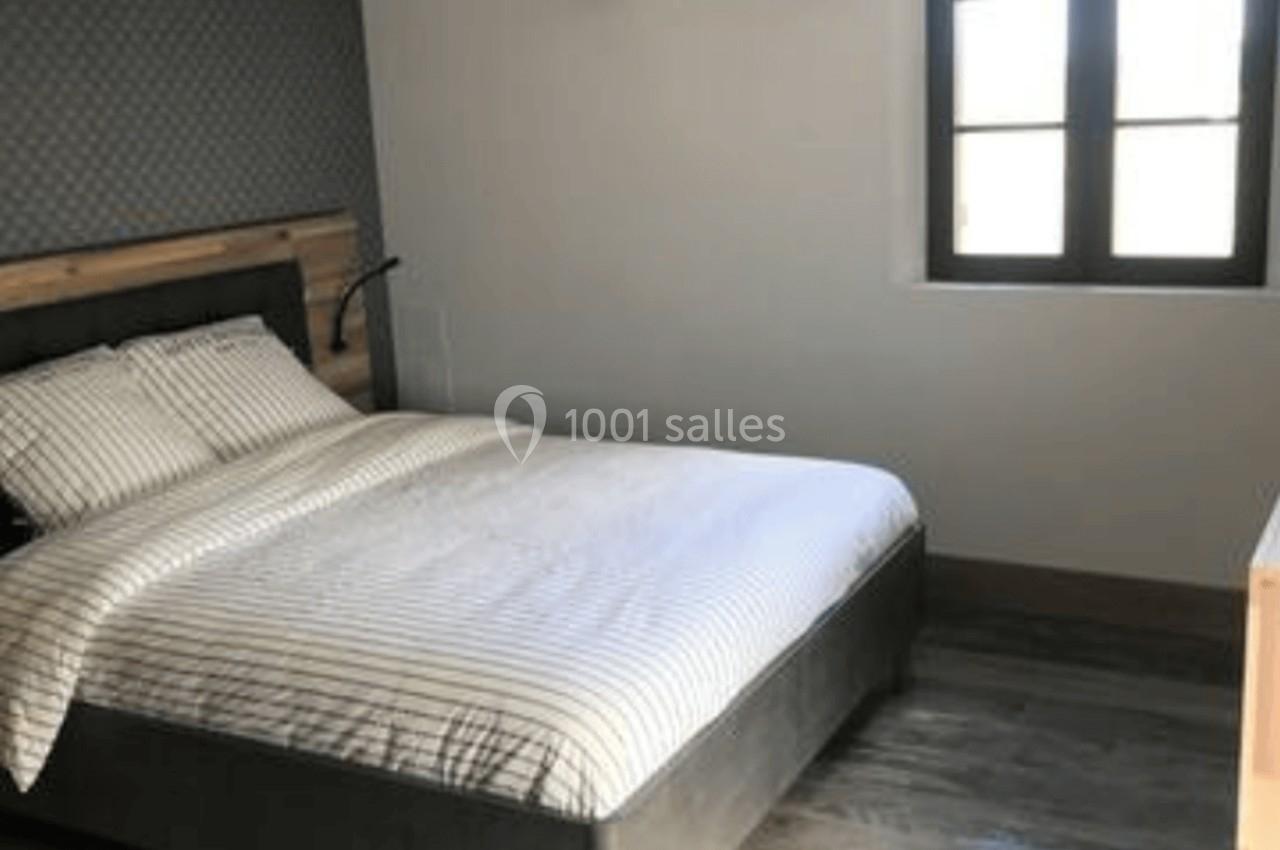Chambre lumineuse avec lit double, parquet en bois, poutres apparentes, mur gris et fenêtre à double battant.