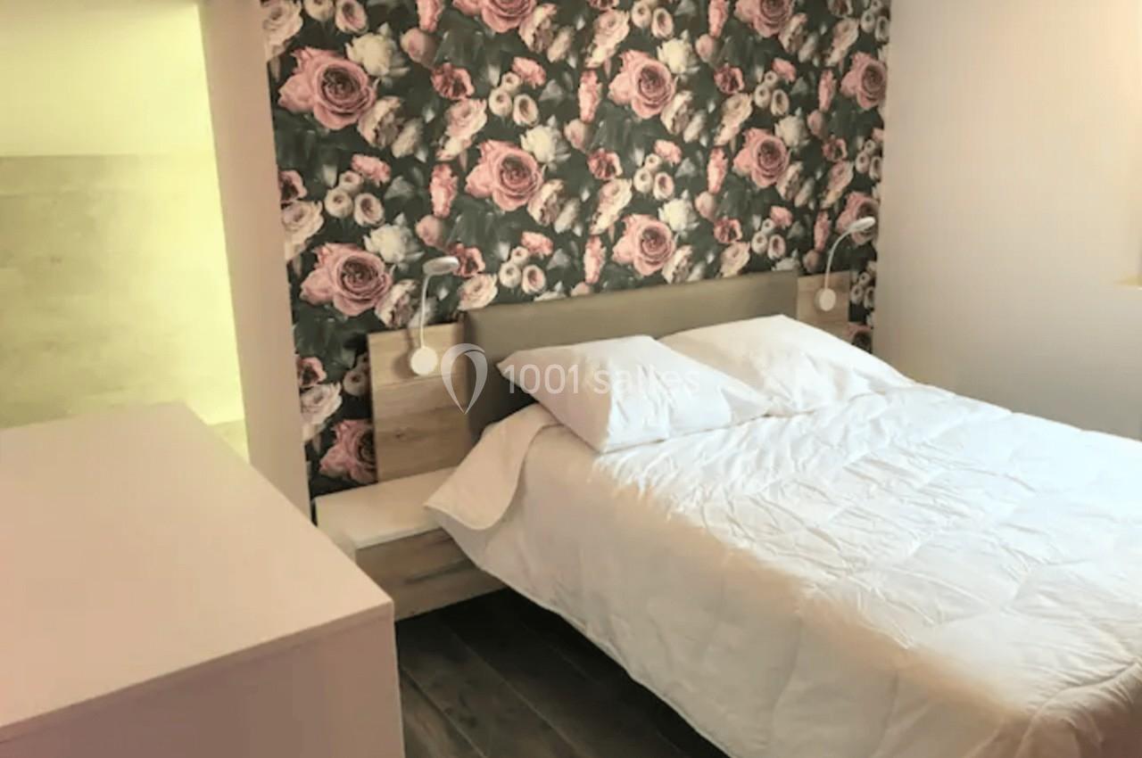Chambre avec lit double, mur décoré de papier peint floral, chevets avec lampes et sol en parquet foncé.