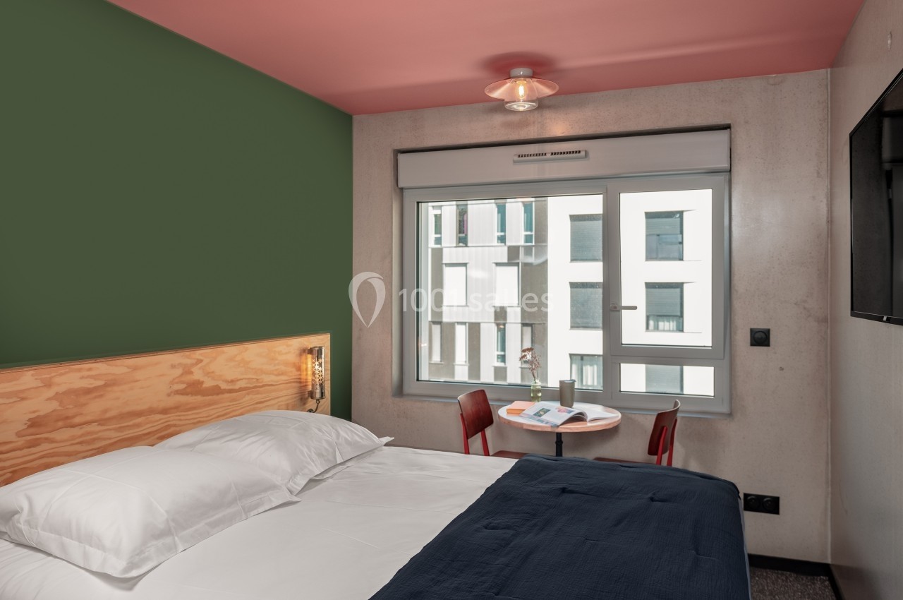 Chambre d'hôtel moderne avec lit double, mur vert, petite table ronde près de la fenêtre et vue sur des bâtiments voisins.