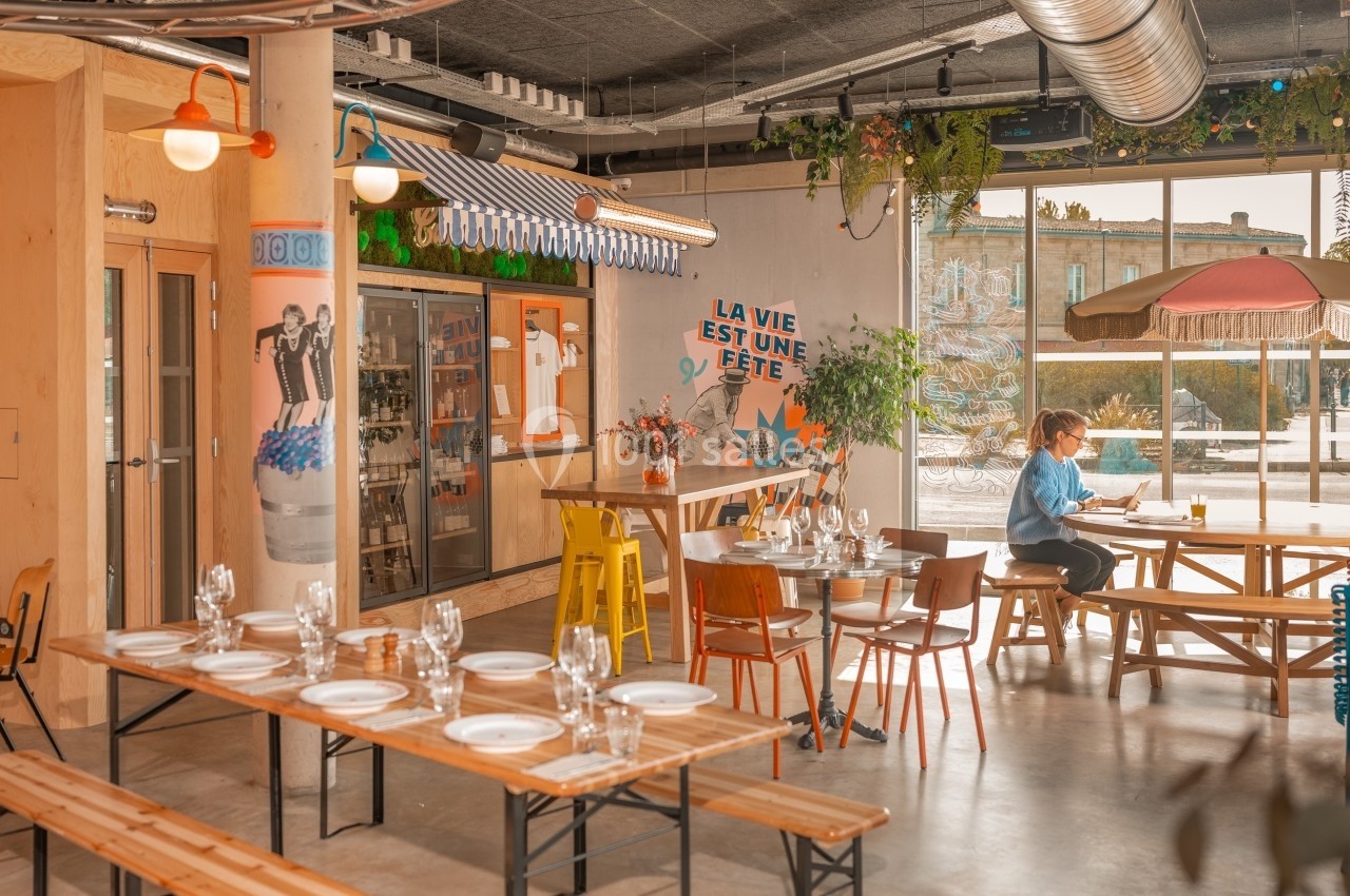Salle de restaurant lumineuse avec tables en bois dressées, décor coloré et une personne assise près d'une baie vitrée.