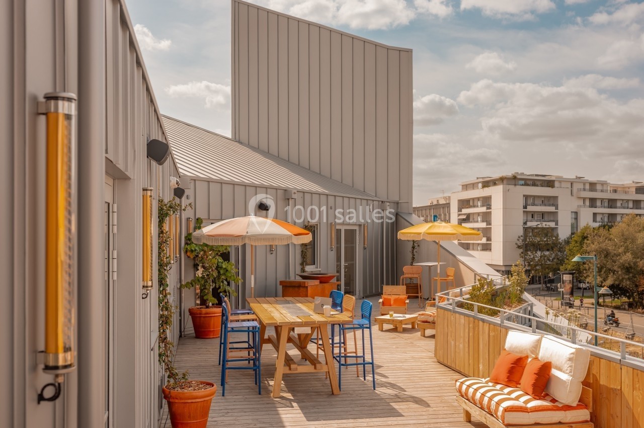 Terrasse en bois avec tables, chaises colorées, parasols, plantes en pot et vue sur des bâtiments résidentiels.