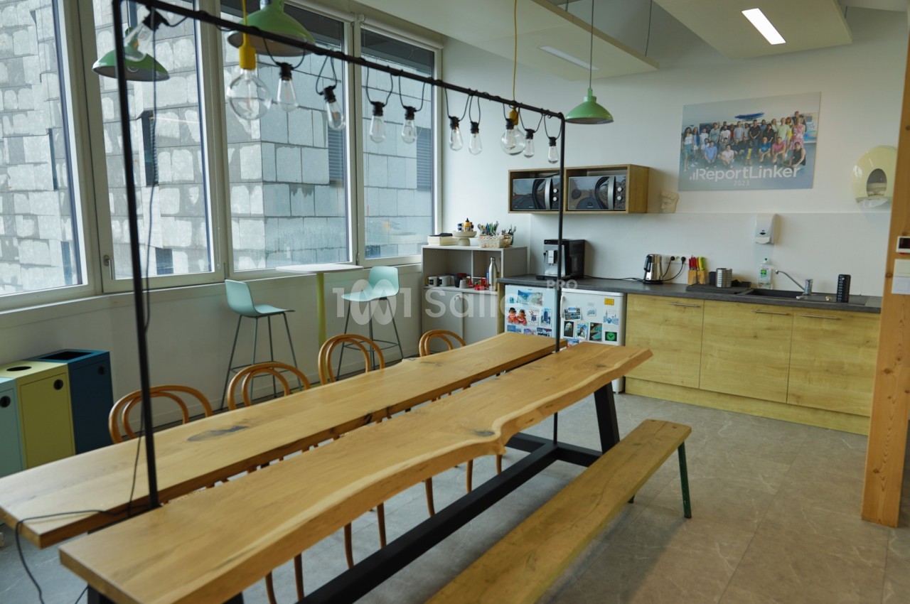 Salle de pause lumineuse avec table en bois, chaises, cuisine équipée et grandes fenêtres donnant sur un mur extérieur.