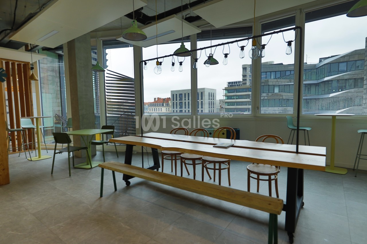 Espace de pause lumineux avec grande table en bois, chaises, tabourets et vue sur des immeubles modernes.