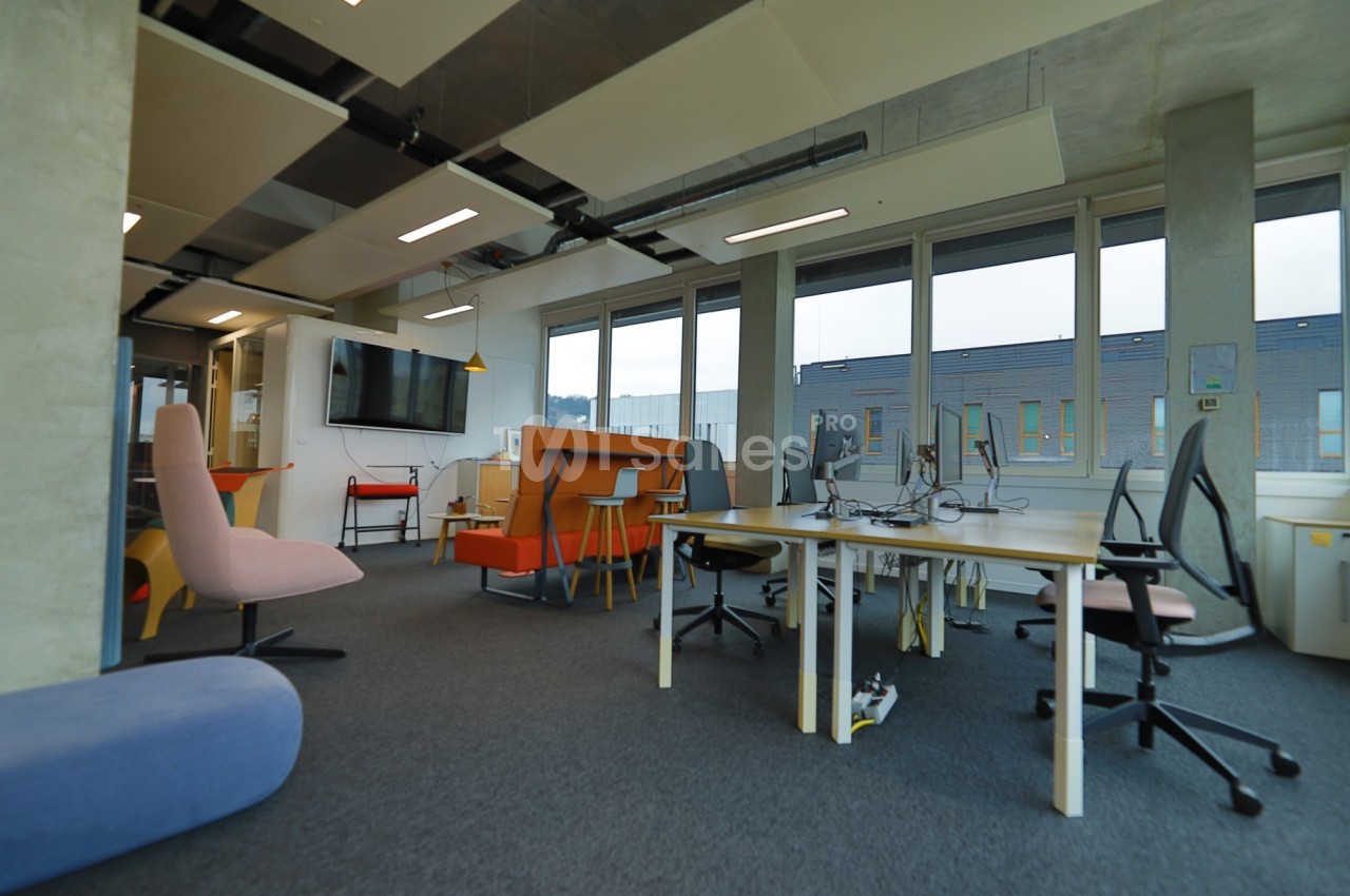 Espace de travail moderne avec bureaux partagés, chaises colorées, grande fenêtre et écran mural.