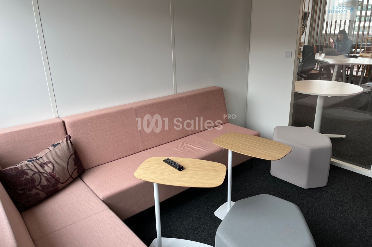 Canapé d'angle rose avec coussin, tables basses en bois clair et poufs gris dans un espace de détente lumineux.