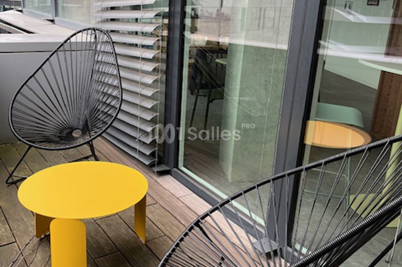 Deux fauteuils noirs et une table jaune sur une terrasse en bois, près de baies vitrées et de stores.