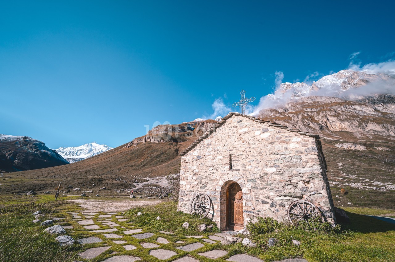 Petite chapelle en pierre avec une croix sur le toit, entourée de montagnes sous un ciel bleu dégagé.