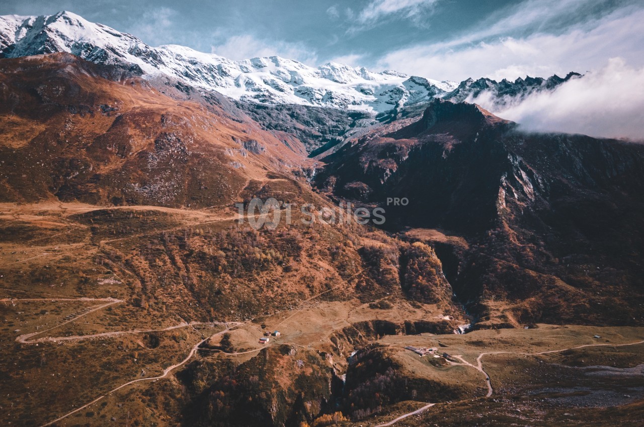 Paysage de montagne avec sommets enneigés, vallées verdoyantes et chemins sinueux sous un ciel partiellement nuageux.