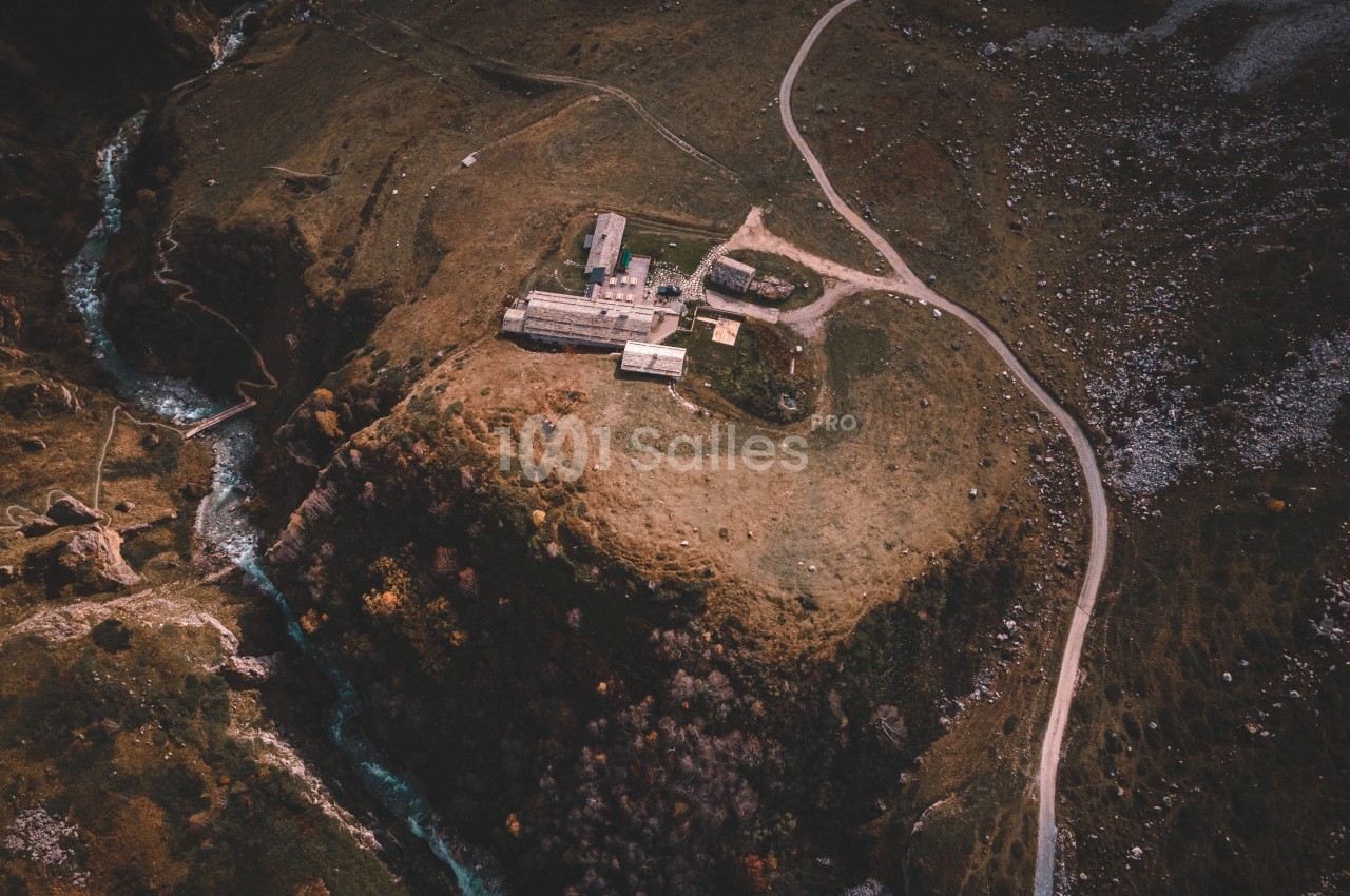 Vue aérienne d'un bâtiment isolé sur une colline entourée de sentiers, d'un ruisseau et de paysages naturels.
