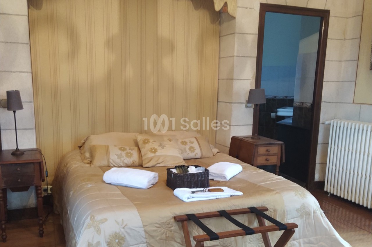 Lit double avec couvre-lit beige, deux tables de chevet, une corbeille d'accueil et une salle de bain visible en arrière…