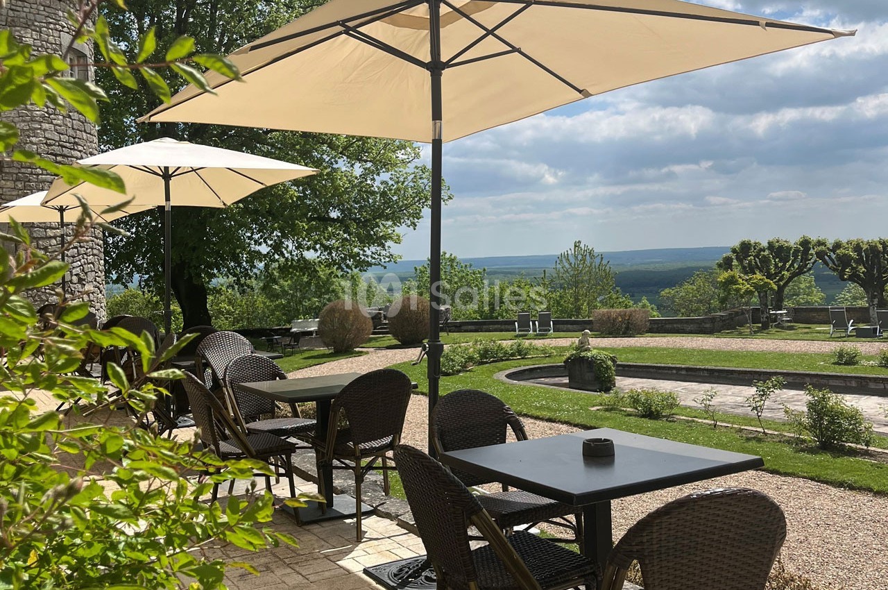 Terrasse avec tables et chaises en osier sous des parasols, offrant une vue dégagée sur un paysage verdoyant.
