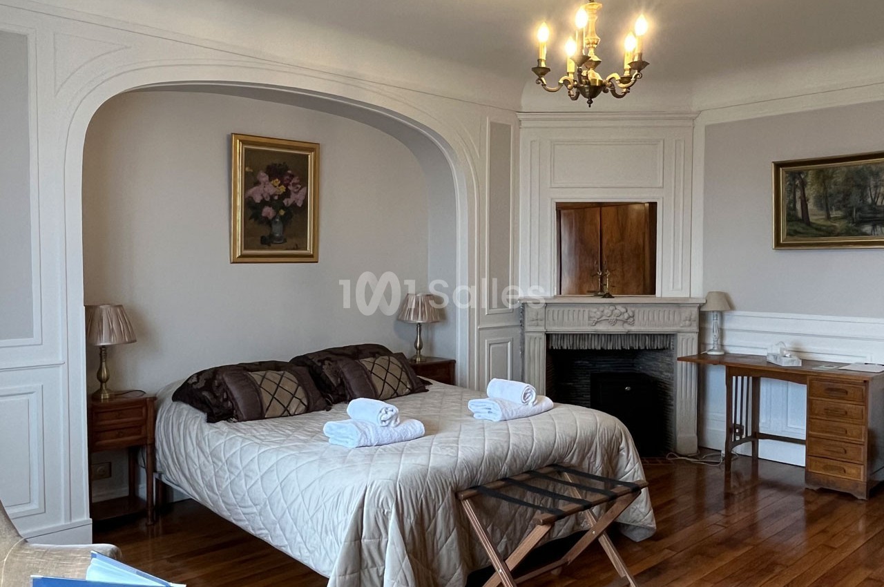 Chambre lumineuse avec lit double, tables de chevet, cheminée décorative, bureau en bois et lustre au plafond.