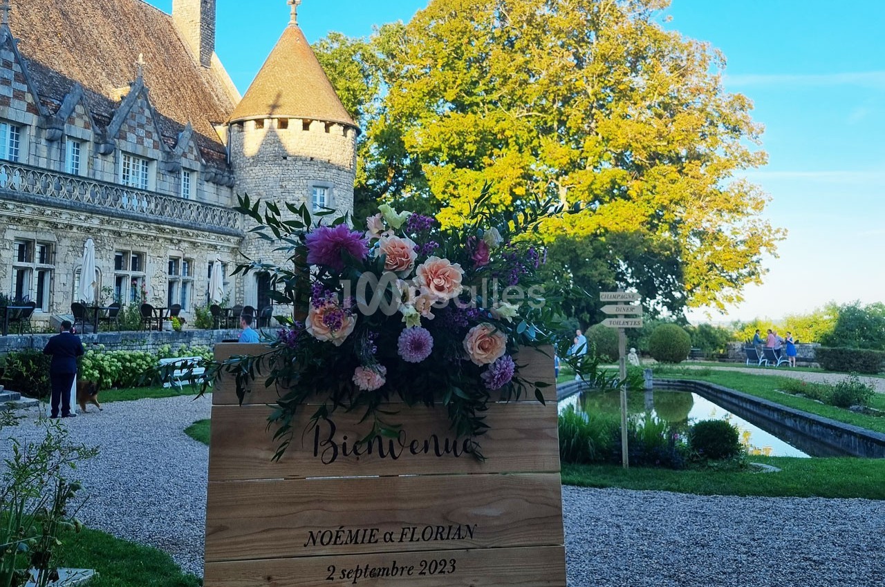 Panneau en bois décoré de fleurs devant un château avec jardin, canal et ciel dégagé en arrière-plan.
