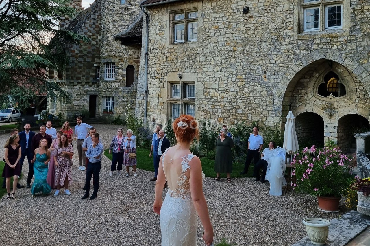 Une mariée en robe blanche fait face à des invités rassemblés dans la cour d'un bâtiment en pierre.
