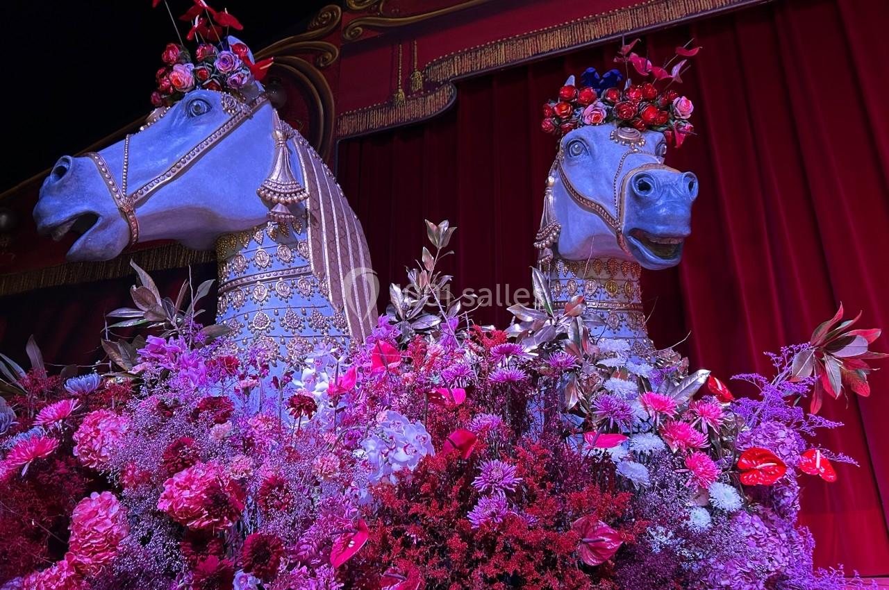Deux statues de chevaux ornées de fleurs et de décorations dorées devant un rideau rouge, entourées de fleurs colorées.