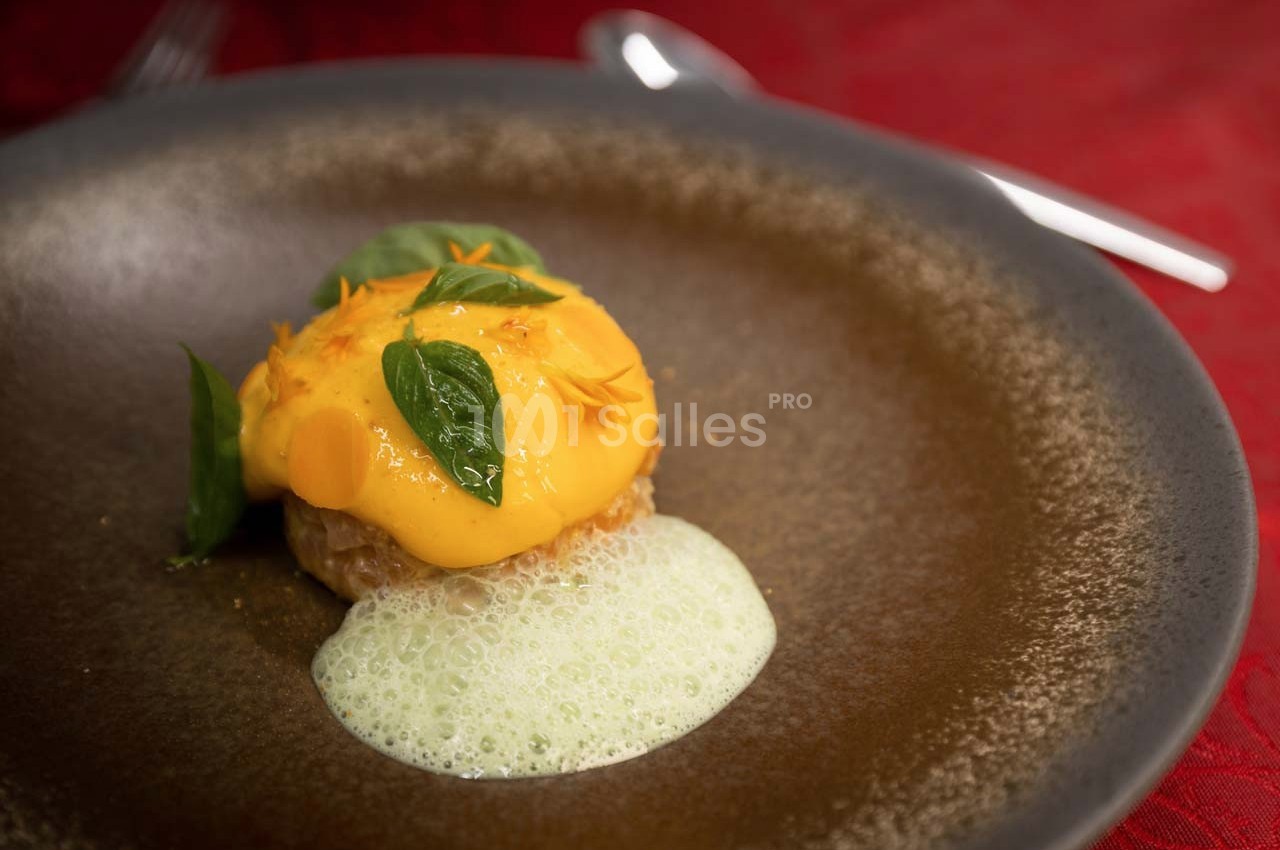 Plat gastronomique composé d'une mousse jaune, de feuilles vertes et d'une émulsion blanche sur une assiette brune.
