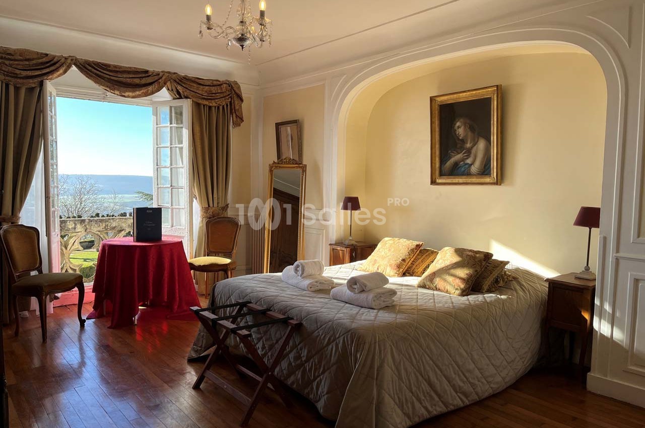 Chambre lumineuse avec lit double, table rouge, tableau au mur et vue sur un jardin et la mer par une porte-fenêtre ouverte.