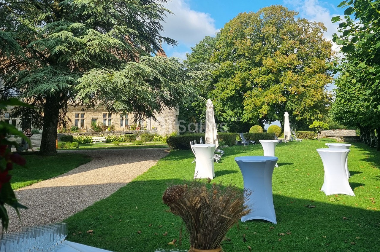 Jardin verdoyant avec tables hautes drapées de blanc, allée gravillonnée et bâtiment en pierre en arrière-plan.