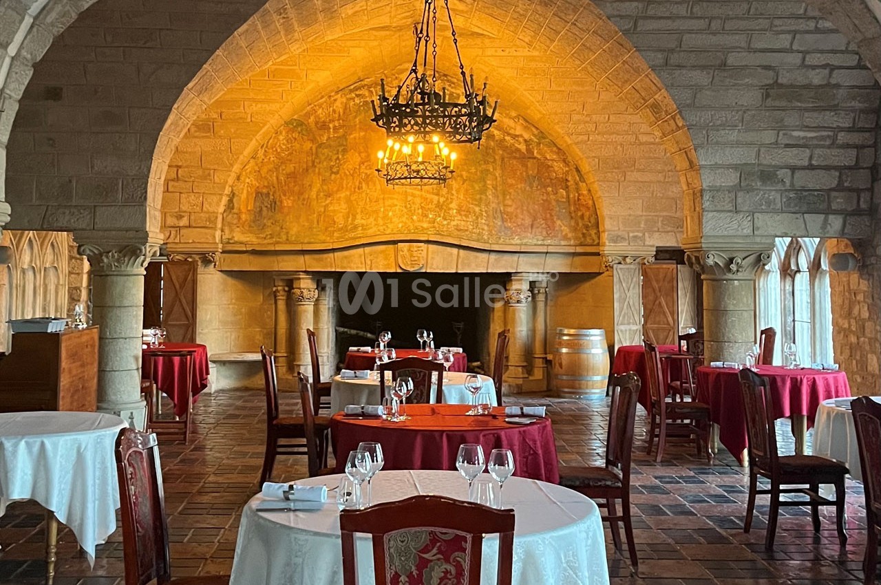 Salle de restaurant au style médiéval avec tables dressées, chandeliers suspendus et fresque murale en arrière-plan.