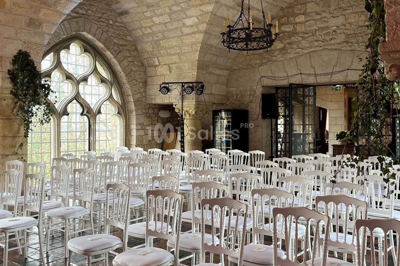 Salle en pierre avec des rangées de chaises blanches alignées, éclairée par de grandes fenêtres gothiques.