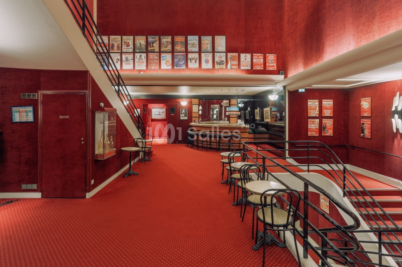 Intérieur d'un théâtre avec moquette rouge, affiches encadrées aux murs, tables et chaises près d'une rampe.
