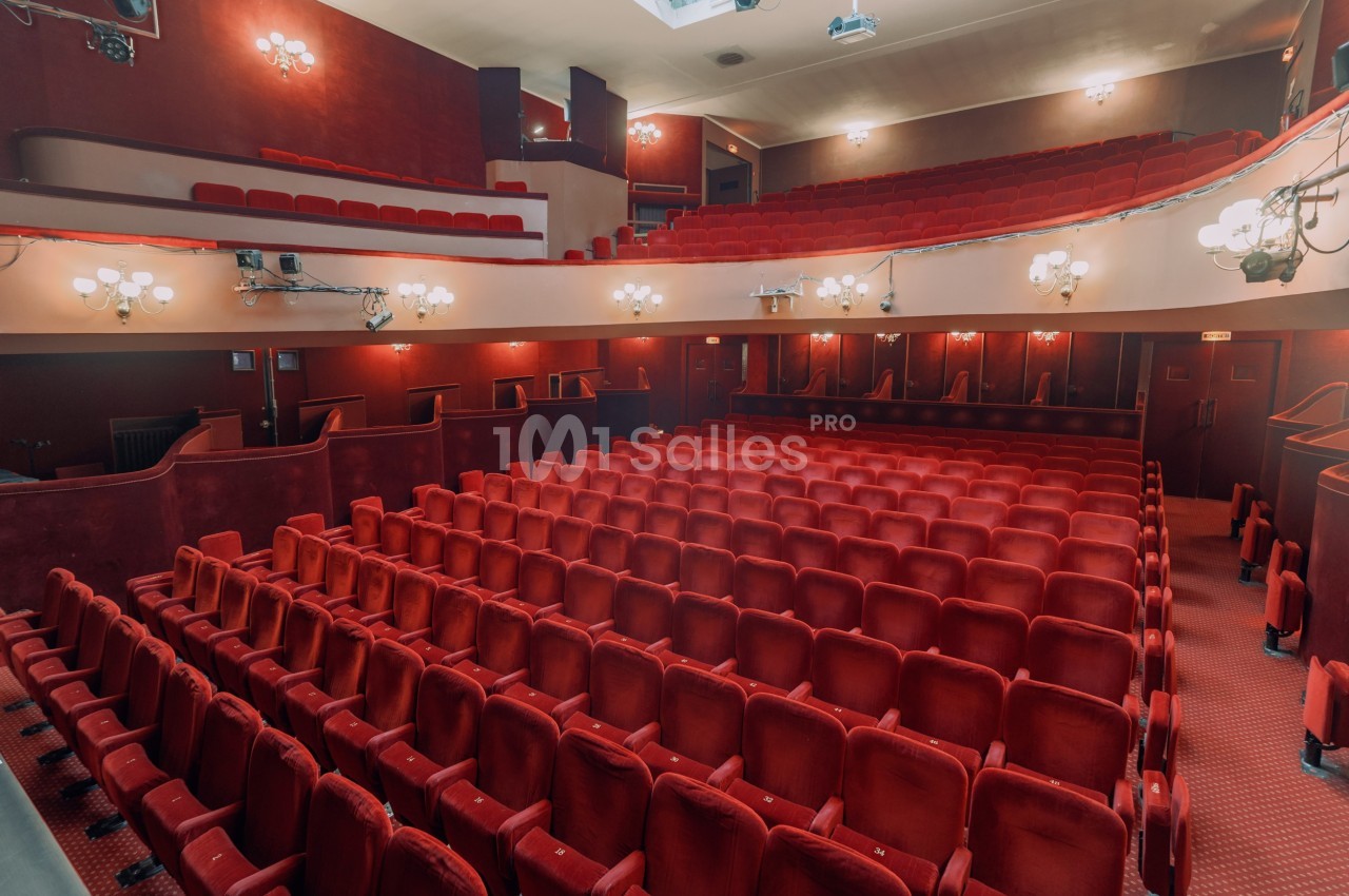 Salle de théâtre vide avec des rangées de fauteuils rouges, éclairage tamisé et balcons supérieurs.