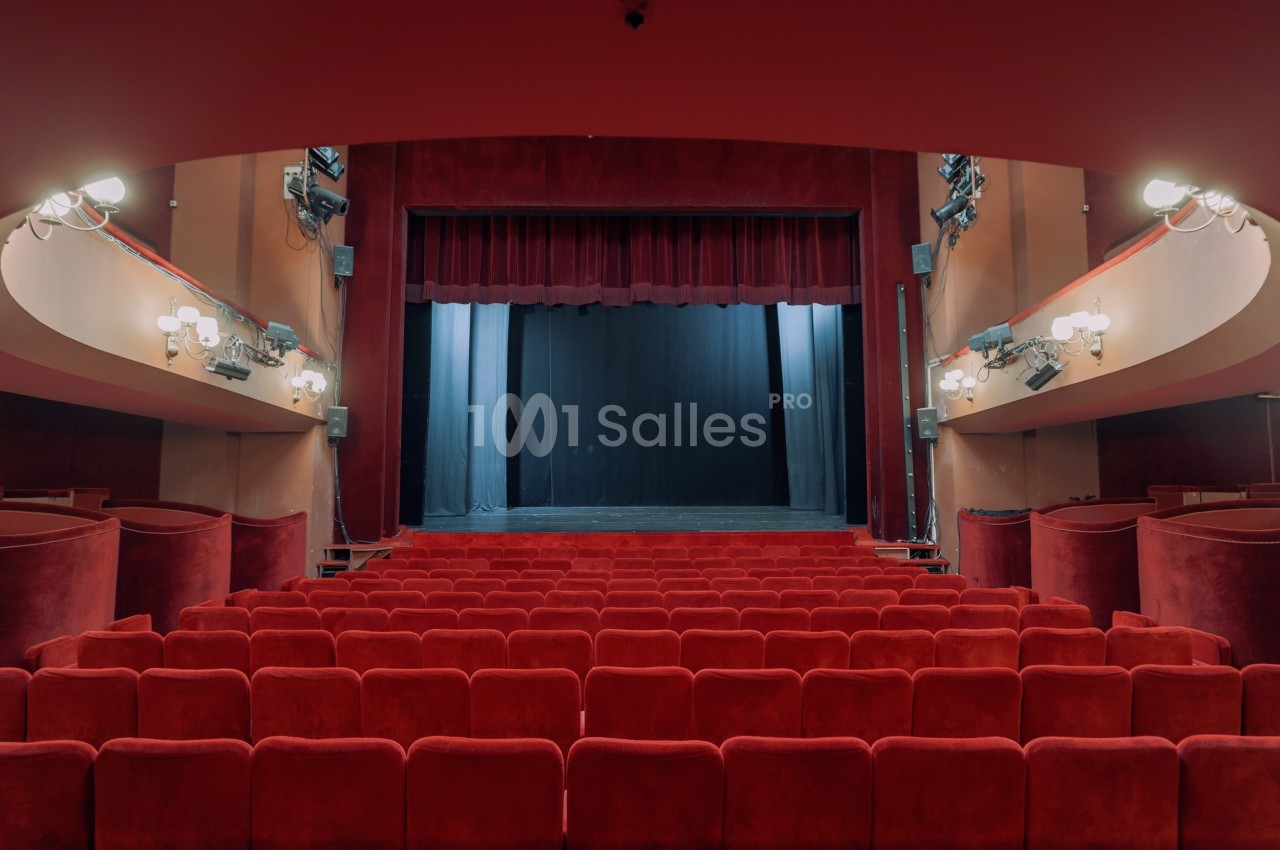Salle de théâtre vide avec des rangées de sièges rouges et une scène encadrée par des rideaux rouges.
