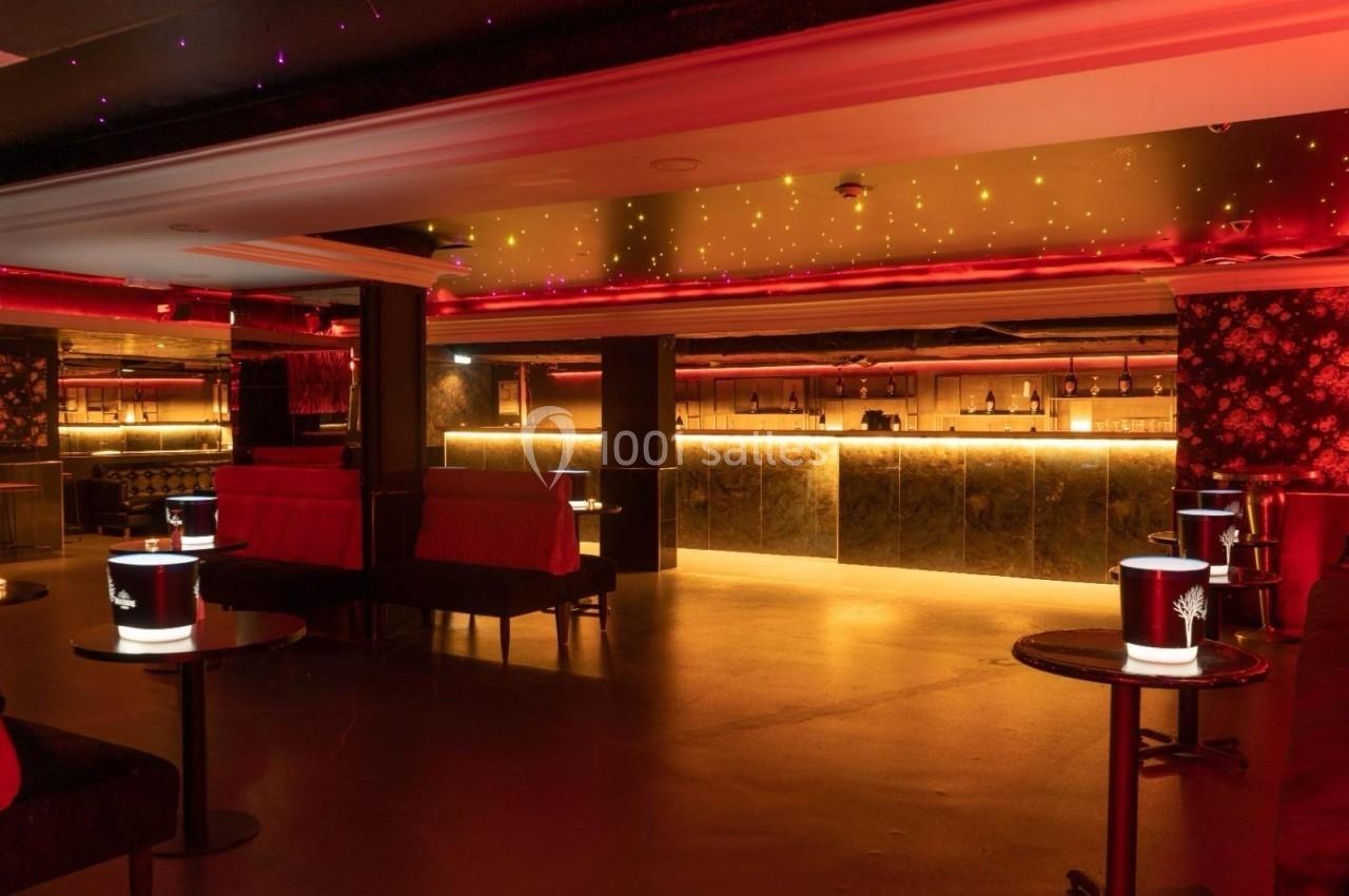 Salle de bar lounge avec éclairage tamisé rouge, banquettes, tables basses et comptoir illuminé en arrière-plan.