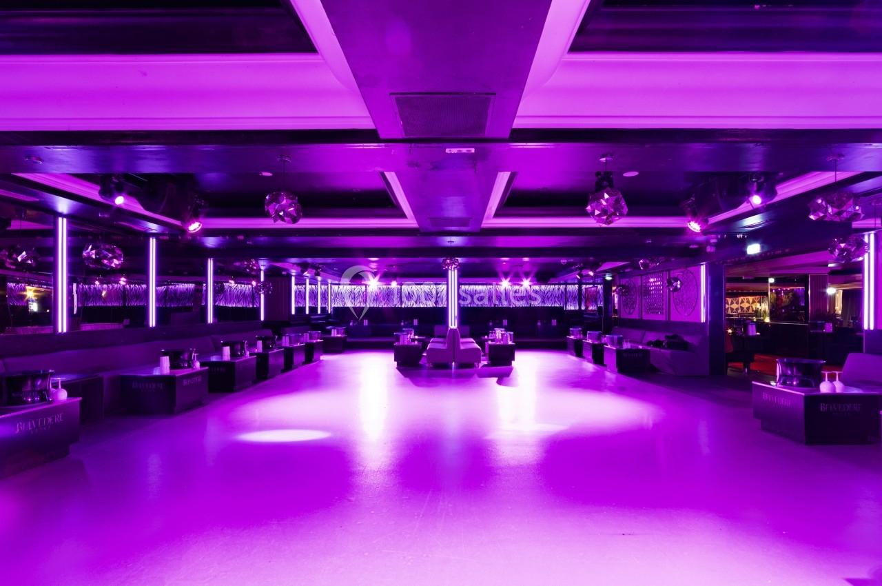 Salle de danse vide éclairée par des lumières violettes, avec banquettes et tables disposées sur les côtés.