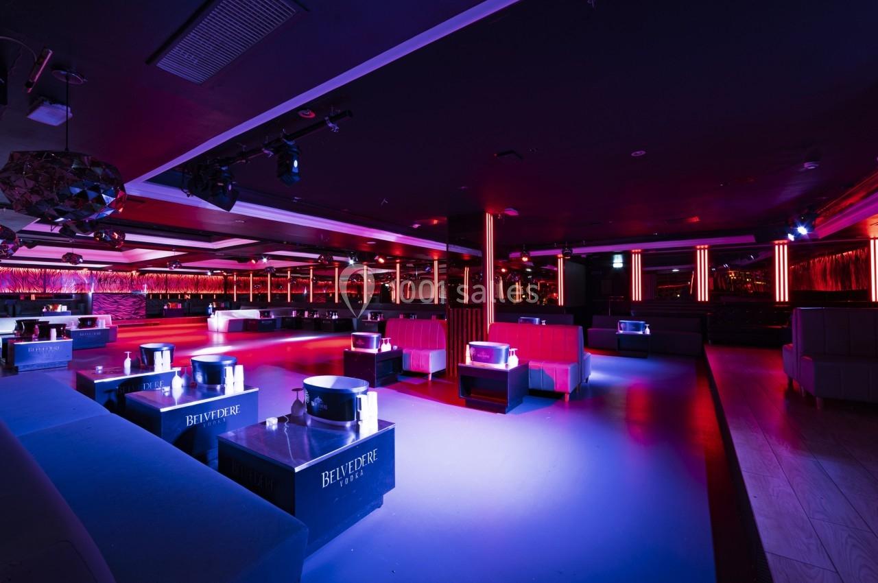 Salle de club nocturne avec éclairage tamisé rouge et bleu, tables basses et banquettes disposées autour d'une piste.