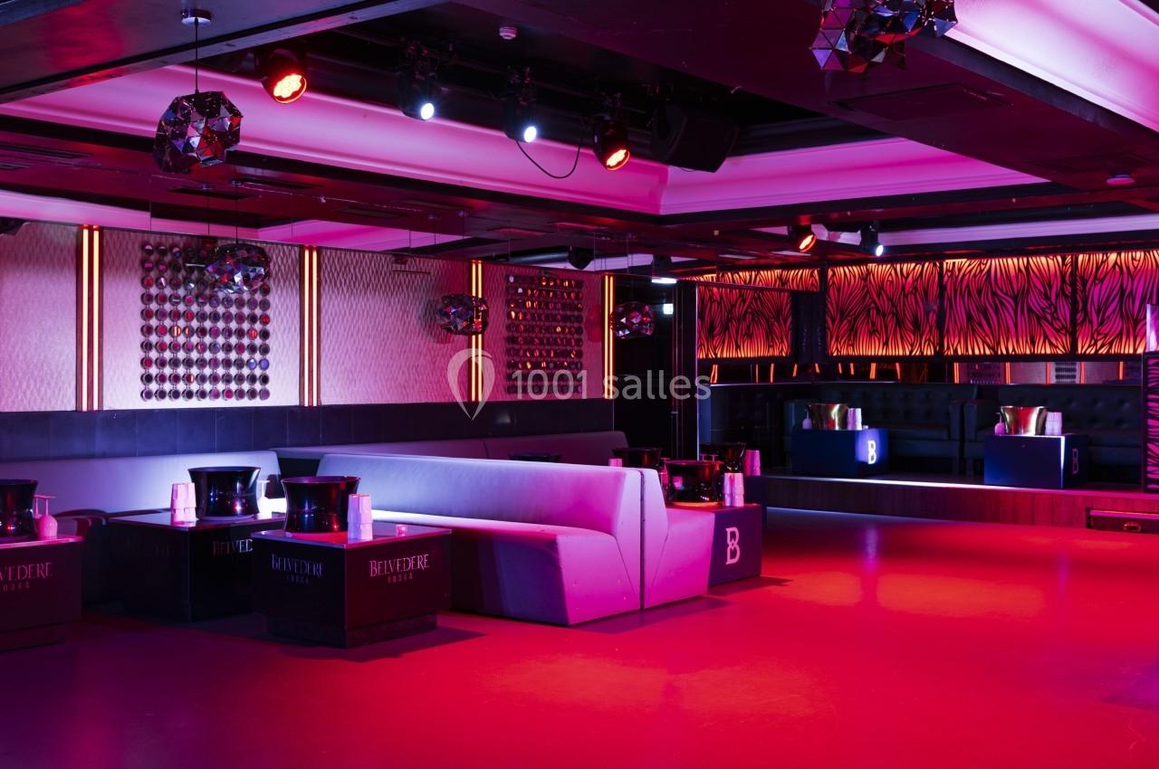Salle de club avec éclairage rouge et violet, banquettes grises, tables basses et décorations murales modernes.