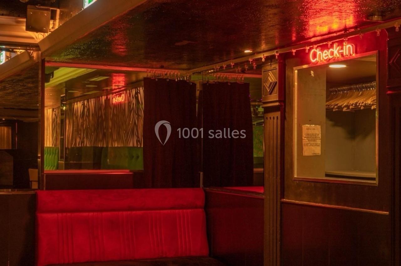 Intérieur d'un espace sombre avec banquette rouge, rideaux noirs et un panneau lumineux ’Check-in’.